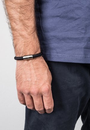 Black braided bracelet with a silver metal clasp, worn on a wrist, featuring a smooth texture and cylindrical design.
