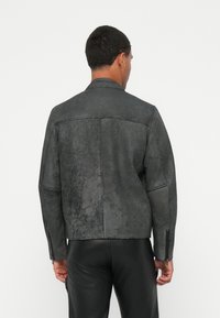 Chaqueta de cuero gris con un acabado texturizado, que presenta un cuello, mangas largas y un detalle de costura en la parte posterior. Los bolsillos con cremallera en la parte delantera no son visibles.