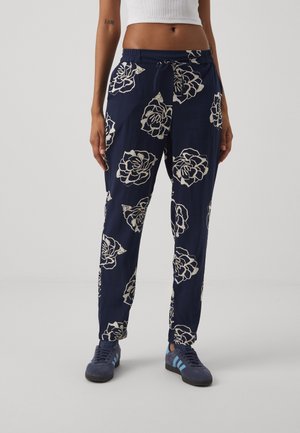 Frau trägt marineblaue Hosen mit großen weißen Blumenmustern, ein weißes kurzes Top und marineblaue Turnschuhe mit hellblauen Streifen.