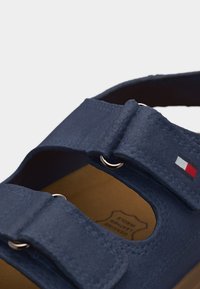 Μπλε δερμάτινο σανδάλι με ρυθμιζόμενους ιμάντες που διαθέτουν μεταλλικούς κρίκους και μικρό λογότυπο με σημαία στο πλάι.