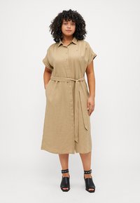 Khakifarbenes Leinenhemdkleid mit kurzen umgeschlagenen Ärmeln, Knopfleiste, Gürtel in der Taille und seitlichen Taschen. Kombiniert mit schwarzen Pumps.
