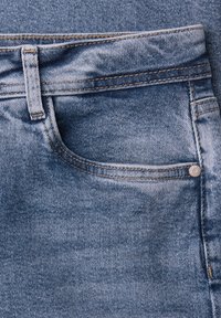 Close-up van de voorkant van een blauwe spijkerbroek met zak, riemlus, zichtbare stiknaden en metalen klinknagel op een gestructureerde stof.