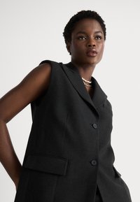 Blazer senza maniche nero con colletto a rever, due tasche frontali e chiusura a singolo bottone, realizzato in un tessuto liscio e strutturato.