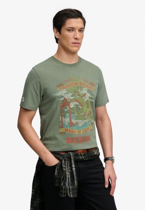 Mann in grünem Grafik-T-Shirt mit Drachendesign, kariertes Hemd um die Taille gebunden, schwarze Hose und Armbanduhr, steht mit einer Hand in der Tasche.