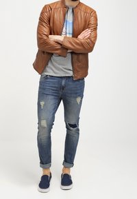 Chaqueta de cuero marrón sobre una camiseta gráfica gris, combinada con jeans azules desgastados y zapatos náuticos azul marino. Ajuste relajado, con rasgaduras visibles en los jeans.