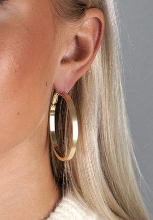 Zbliżenie ucha osoby o długich blond włosach, noszącej duże, gładkie, złote kolczyki koła na szarym tle.