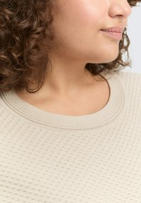 Beige gebreide top met een gestructureerd patroon, geribbelde ronde halslijn en subtiele details, die zacht textiel in een close-up weergave toont.