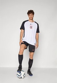 Fiatal férfi fekete-fehér focimezben, egyik lábával egy fehér-kék focilabdán áll, egyszínű háttér előtt.