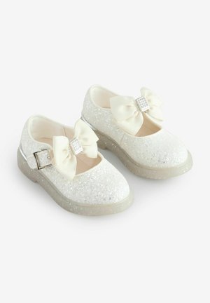 Chaussures blanches pailletées avec un bout arrondi, une semelle transparente, une boucle réglable et deux grands nœuds crème ornés de strass.