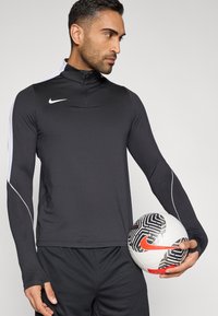 Μαύρο αθλητικό μπλουζάκι με μακριά μανίκια και ψηλό γιακά, με λευκές πλευρικές λεπτομέρειες, φορεμένο από άτομο που κρατάει μια ποδοσφαιρική μπάλα με σχέδια.