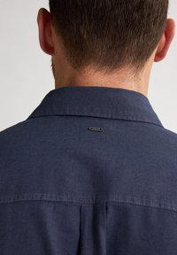 Laivastonsininen paita, jossa on rakenteellinen kaulus, hienovarainen ommel ja pieni logotagi takana. Kangas vaikuttaa sileältä ja kevyeltä.