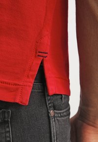 Vue latérale d'une personne portant un polo rouge rentré à l'extérieur d'un jean noir, montrant la fente latérale et les détails de couture du polo.