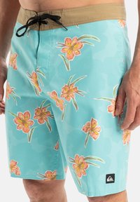 Tyrkysové plavky s kvetinovým vzorom, na ktorých sú oranžové kvety a zelené listy. Hnedý pás a podrobnosti šnúrky. Štítok s logom na leme.