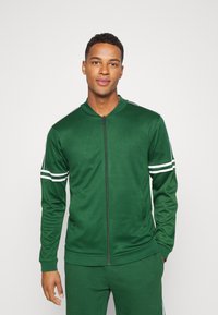 Grüne sportliche Zip-Jacke aus glattem Stoff. Mit weißen Streifen an den Ärmeln und Stehkragen. Taschen auf beiden Seiten.