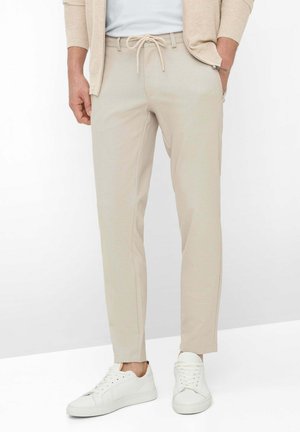 Beige slim-fit broek met trekkoorden in de taille en zakken, gedragen met witte sneakers en een beige jack met ritssluiting.