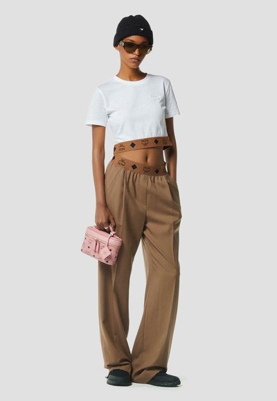 Femme portant un t-shirt blanc court, un pantalon marron taille haute avec une ceinture de marque, un bonnet noir, des lunettes de soleil, et tenant un petit sac à main rose.