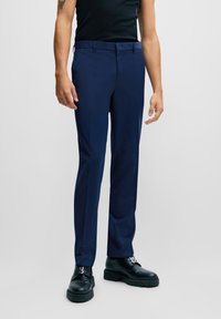 Marineblaue Slim-Fit-Hosen mit Gürtelschlaufen und seitlichen Taschen, aus glattem Stoff gefertigt. Getragen mit einem schwarzen Tanktop und schwarzen klobigen Schuhen.