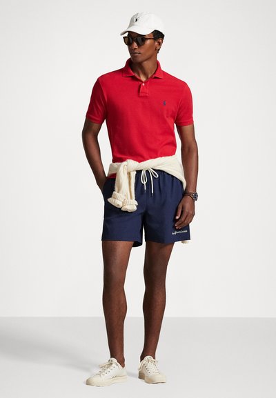 Κόκκινο πόλο μπλουζάκι, ναυτικές βερμούδες, λευκά αθλητικά παπούτσια και λευκό καπέλο. Ζακέτα δεμένη στη μέση, γυαλιά ηλίου φορεμένα στο πρόσωπο. Χαλαρή καλοκαιρινή εμφάνιση.