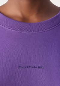 Lähikuva henkilöstä, joka pukeutuu violettiin pyöreäkaulaiseen paitaan, jonka rinnassa on pienellä tummalla tekstillä brodeerattu "Marc O'Polo DENIM".