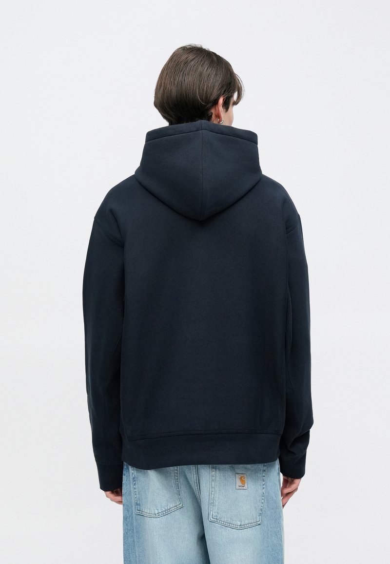 Marineblauwe hoodie met een grote capuchon, gemaakt van zacht materiaal. Heeft lange mouwen en een ontspannen pasvorm, gecombineerd met lichtgewassen spijkerbroek.