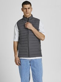Graue gepolsterte Weste mit hohem Kragen, Reißverschluss und gestepptem Design, getragen über einem hellgrauen T-Shirt, kombiniert mit lockeren blauen Jeans.