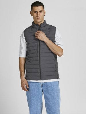 Graue gepolsterte Weste mit hohem Kragen, Reißverschluss und gestepptem Design, getragen über einem hellgrauen T-Shirt, kombiniert mit lockeren blauen Jeans.