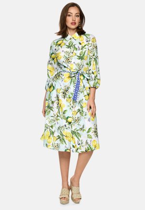 Frau trägt ein weißes Midikleid mit gelbem Zitronen- und grünem Blattmuster, Puffärmeln, blau gemustertem Taillenband und beigen, gewebten Plateausandalen.