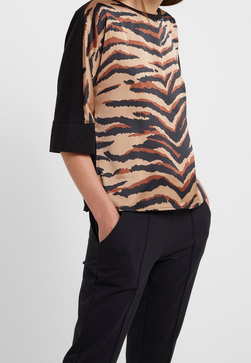 Femme portant un haut beige et noir à rayures tigre à manches courtes avec des manches noires unies et un pantalon noir, les mains dans les poches.