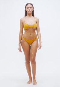 Σετ μπικίνι μουσταρδί με λεπτές τιράντες, με απλό, μινιμαλιστικό σχέδιο. Το υλικό φαίνεται λείο και ελαστικό.