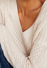 Jasno beżowy sweter z plecionki z dekoltem w kształcie litery V, z fakturowanymi wzorami i ściągaczami na mankietach, swobodnie opadający na ramionach osoby.