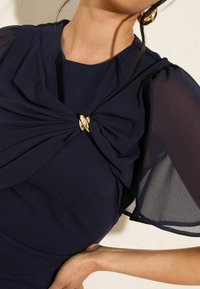 Laivastonsininen mekko, jossa on tekstuuri, kukkakaulusyksityiskohta olkapäällä, kiinnitetty kultasävyiseltä soljella ja läpikuultavat puffihihat.