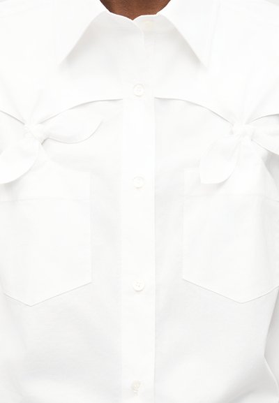 Chemise blanche boutonnée avec deux poches avant et des nœuds en tissu noués près des épaules de chaque côté.