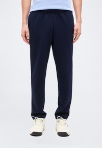 Άτομο που φοράει μπλε σκούρο φόρμας και λευκά αθλητικά παπούτσια, στέκεται μπροστά σε ένα απλό ανοιχτό φόντο.