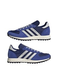 Kék velúrból készült sportcipők fehér talppal. Az oldalán három ezüstcsík található, és texturált orr része van. Szürke fűzőkkel rendelkezik.