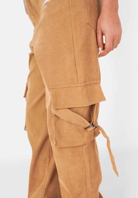 Bruine corduroy cargobroek met flapzakken, verstelbare banden en een gestructureerd uiterlijk. De stof heeft verticale ribbels en een zachte uitstraling.