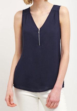 Mouwloze marineblauwe blouse met een ritssluiting aan de voorkant en getextureerde schouderpanelen, gedragen met witte broek.