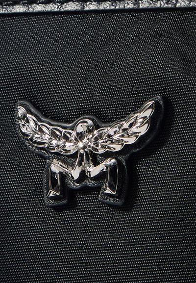 Fényes ezüst absztrakt embléma, szárnyra emlékeztető levelekkel és hajlított lábakkal, texturált fekete szövet háttér előtt.