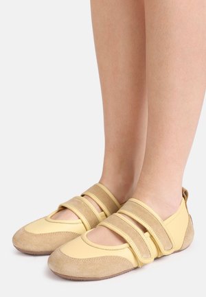 Jalad, mis kannavad kollaka-beeži värvi Mary Jane stiilis kingi, millel on kaks laia reguleeritavat rihma ja tekstuuriga kummist tallad.