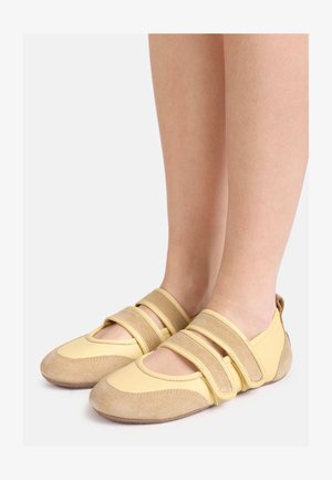 Πόδια που φορούν κίτρινα μπεζ παπούτσια στυλ Mary Jane με δύο φαρδιές ρυθμιζόμενες λουρίδες και υφή από λαστιχένια σόλα.