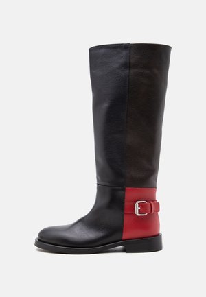 Botte haute en cuir avec tige noire, zone de talon rouge avec boucle argentée et semelle basse noire, montrée de profil sur fond blanc.