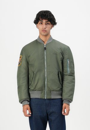 Olijfgroene bomberjack met grijze gebreide manchetten en zoom, met een logo-patch op de linkermouw en een ritszak op de arm.