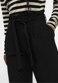 Pantalons noirs ajustés avec une taille large et un nœud à nouer. Caractérisés par des plis et des poches latérales. Portés avec un pull rayé noir et blanc.