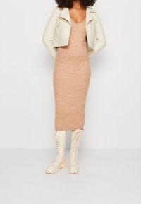 Robe en tricot côtelé beige avec une silhouette ajustée, portée avec une veste en cuir blanche courte et des bottes à lacets crèmes jusqu'aux genoux.