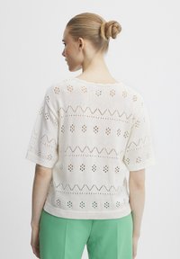 Strickpullover in Creme mit Lochmustern, kurzen Ärmeln und einem gewellten Ausschnitt, kombiniert mit grünen Hosen.
