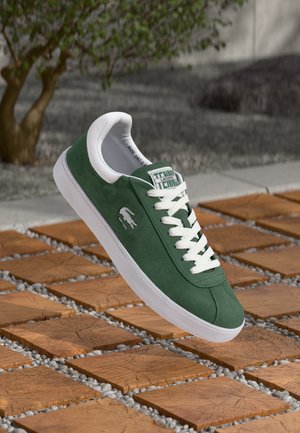 Sneaker verde in suede con dettagli bianchi, dotata di un'etichetta sulla linguetta verde e bianca, lacci bianchi e suola in gomma bianca, sospesa sopra piastrelle di legno.