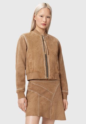 Vrouw met lang blond haar die een bruine suède jas met rits en een bijpassende gestikte suède rok draagt tegen een effen achtergrond.