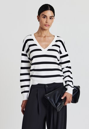 Woman wearing a black and white striped V-neck sweater and black wide-leg trousers holding a large black leather clutch bag.