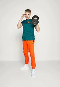 Jauns vīrietis tirkīza krāsas T-kreklā un spilgti oranžās treniņbiksēs paceļ hanteli ar vienu roku, ar otru roku sveicina, stāvot uz vienkārša fona.