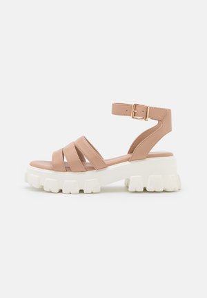 Sandale beige épaisse avec trois brides avant, bride de cheville avec boucle dorée, et semelle épaisse blanche à crampons sur fond uni.