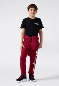 Schwarzes Baumwoll-T-Shirt mit dem Aufdruck "Übung macht den Meister", kombiniert mit roten Jogginghosen mit weißem "Champion"-Logo. Schwarze und rote Sportschuhe.